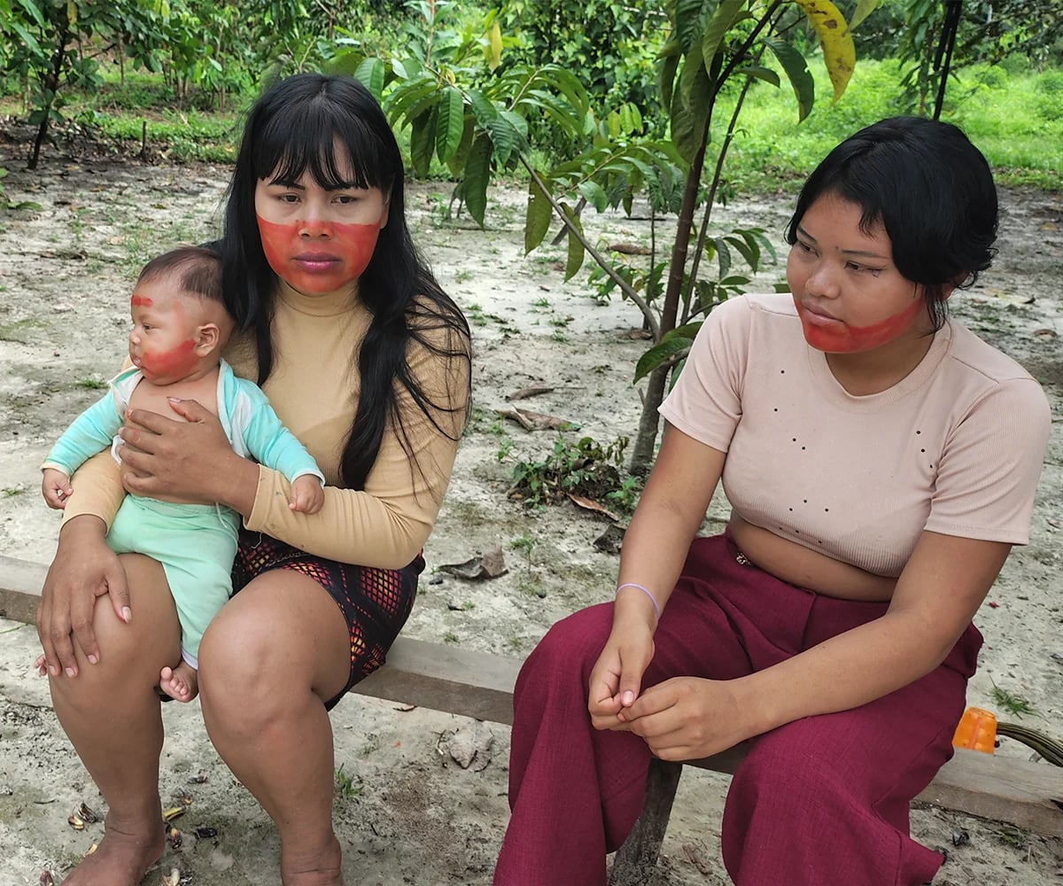 Mulher com um bebê no colo sentada ao lado de outra mulher em uma área arborizada